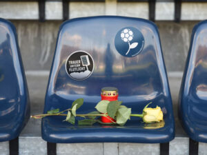 Auf einem blauen Stadionsitz liegt eine gelbe Rose und ein rotes Grablicht steht auf dem Sitz. Auf dem Sitz ist ein Sticker von Trauer und Fußball sowie von Trauer unterm Flutlicht geklebt.