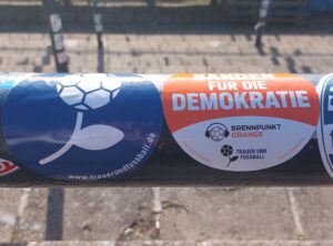 Zwei Sticker im Stehplatzbereich. Einmal die Fußballblume von Trauer und Fußball, einmal der orange Sticker mit Brennpunkt Orange #BildetBandenFürDieDemokratie.